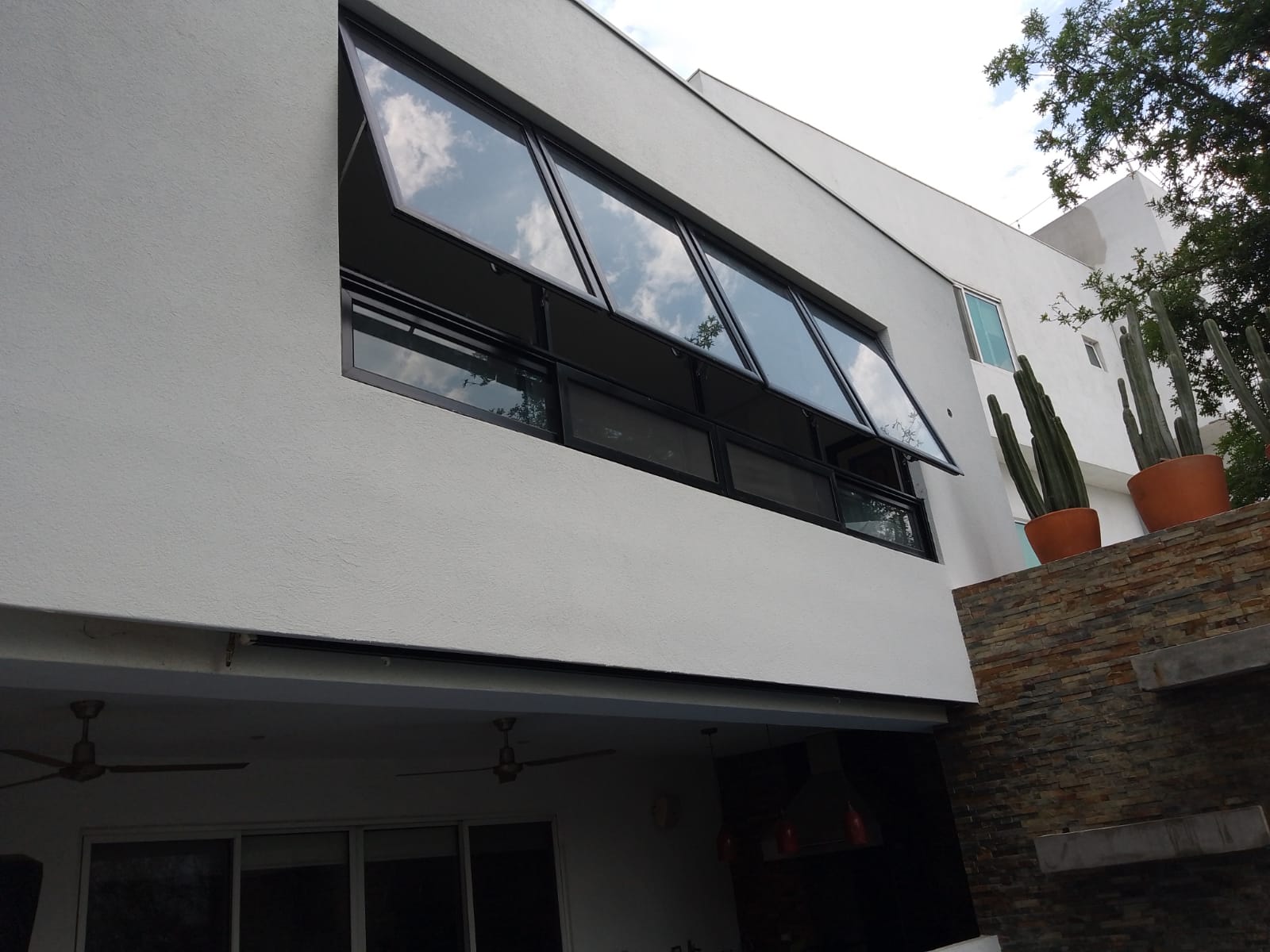 Vista exterior de la fachada con ventanas de proyección tipo taquilla en aluminio negro y cristal reflectivo, sobre área de terraza con cantera.