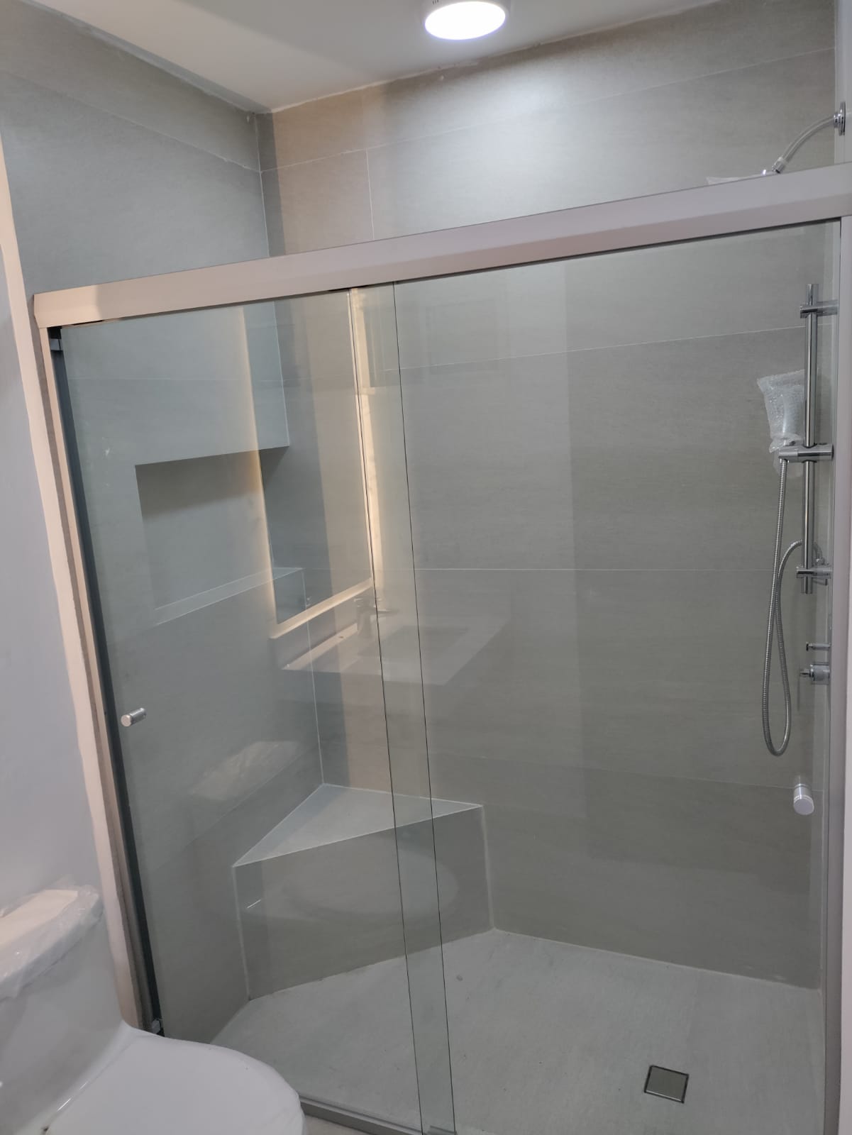 Cancel de baño tipo corredizo con perfil de aluminio y cristal templado, instalado sobre regadera con banca de mármol.