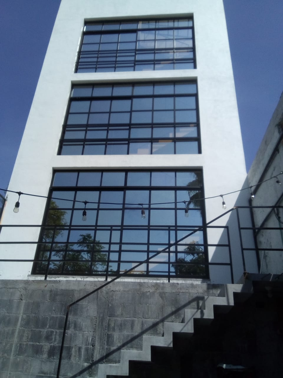 Fachada acristalada de tres niveles con perfilería de aluminio negro y cristal reflectivo, integrada a muros blancos en edificio del centro.