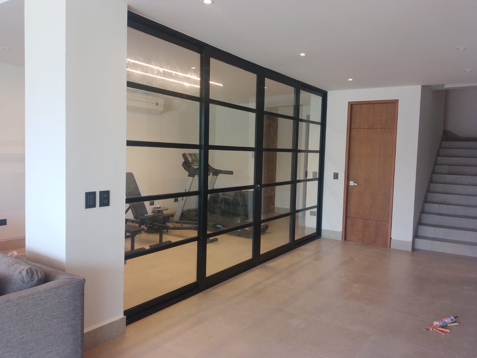 Vista lateral del cancel de aluminio negro con cristal templado y puerta corrediza integrada, dividiendo gimnasio del área social.